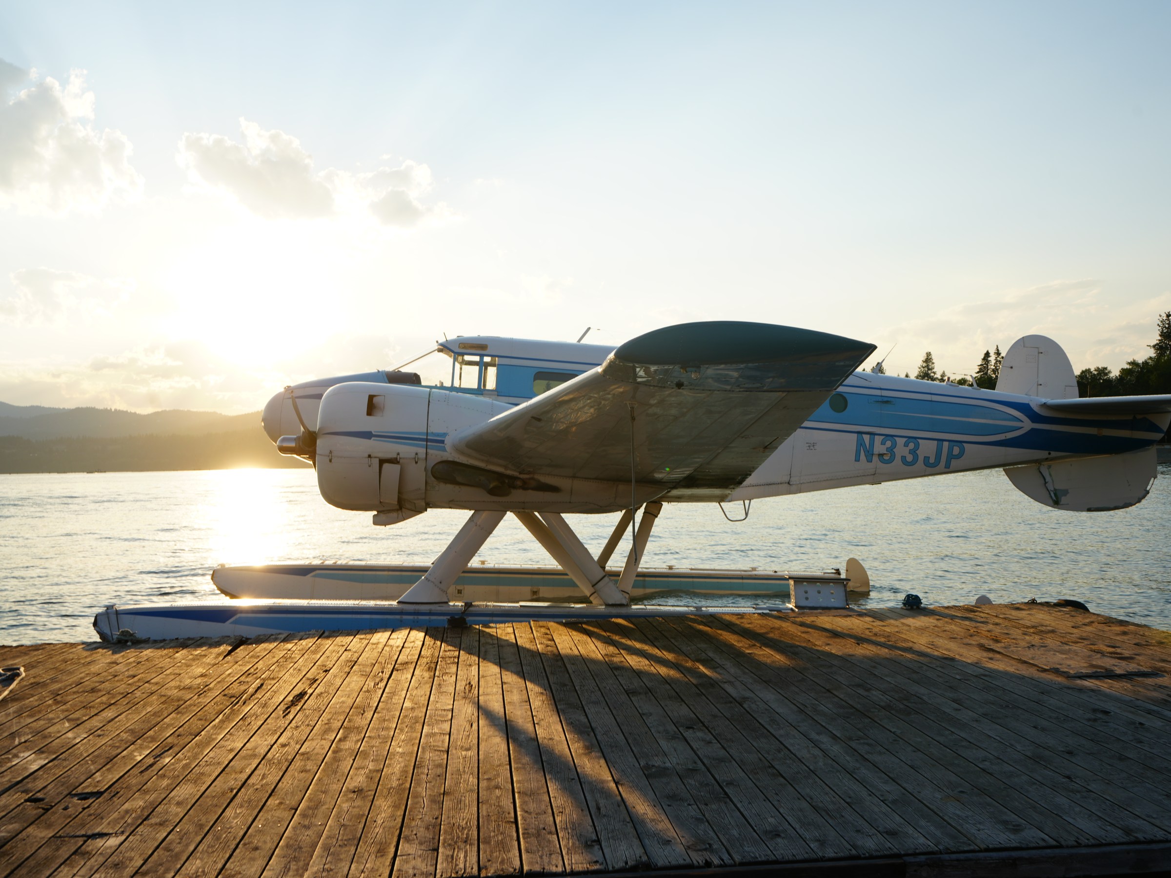 Seaplane docked on wooden pier at sunset, with mountains and trees in background.