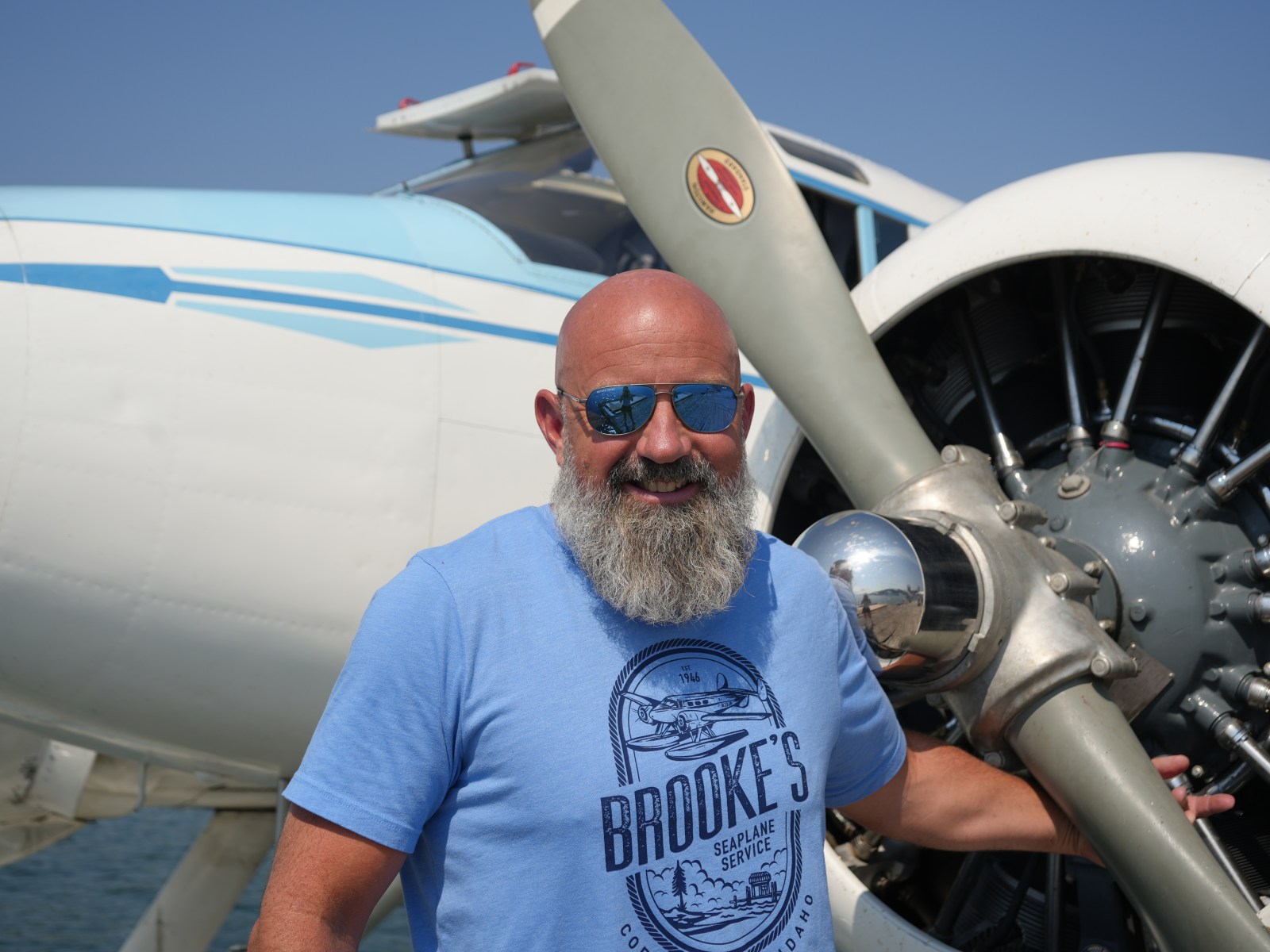 Man with a beard in sunglasses stands by a vintage airplane propeller.