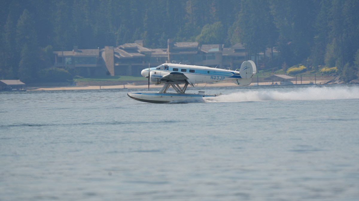 Seaplane taking off from water near a forested shore with buildings in the background.