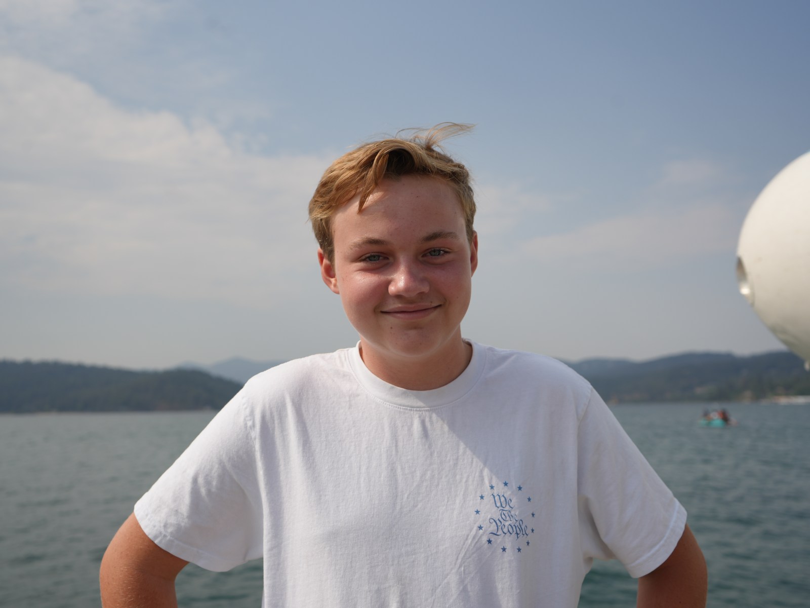 Smiling person in a white shirt standing by a lake on a sunny day.